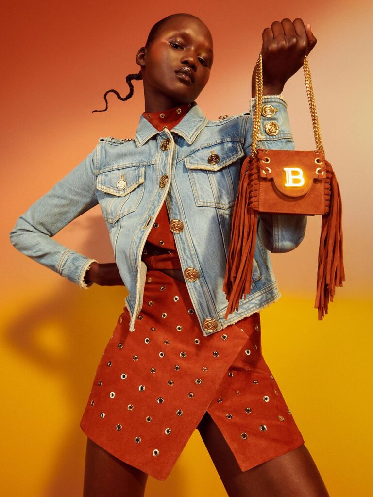 Woman posing in jean jacket and suede skirt, holidng up a fringe balmain bag.